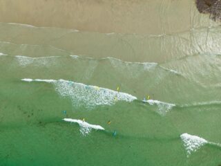 Overhead shot of small waves breaking along a sandy shoreline, with surfers in the water adding movement and energy to the expansive coastal scene