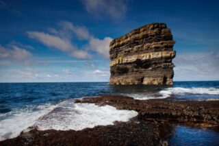 Layered rock sea stack standing offshore against a deep blue sea, with foaming waves and low coastal rocks adding texture and movement to the scene